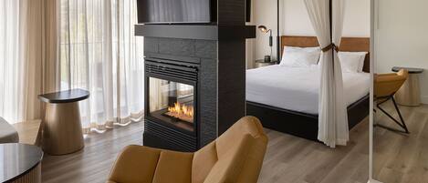 A modern hotel room with a large bed, a fireplace, a sofa, and a dining area.