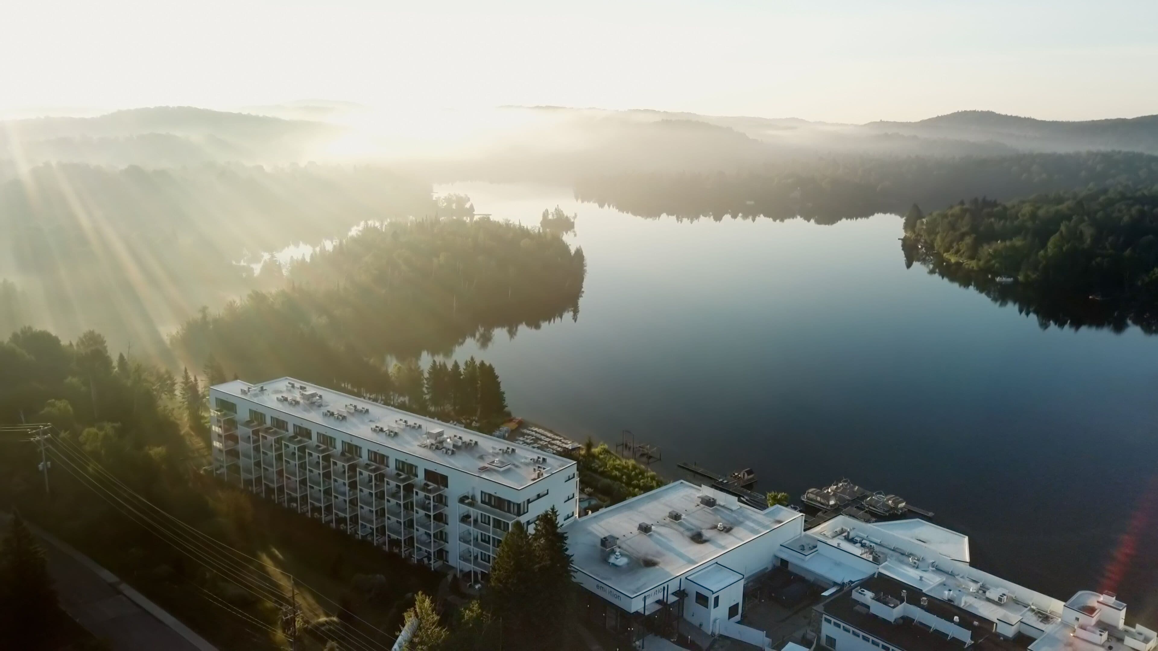 An aerial view of a modern hotel with multiple buildings, surrounded by a serene lake and forested hills.
