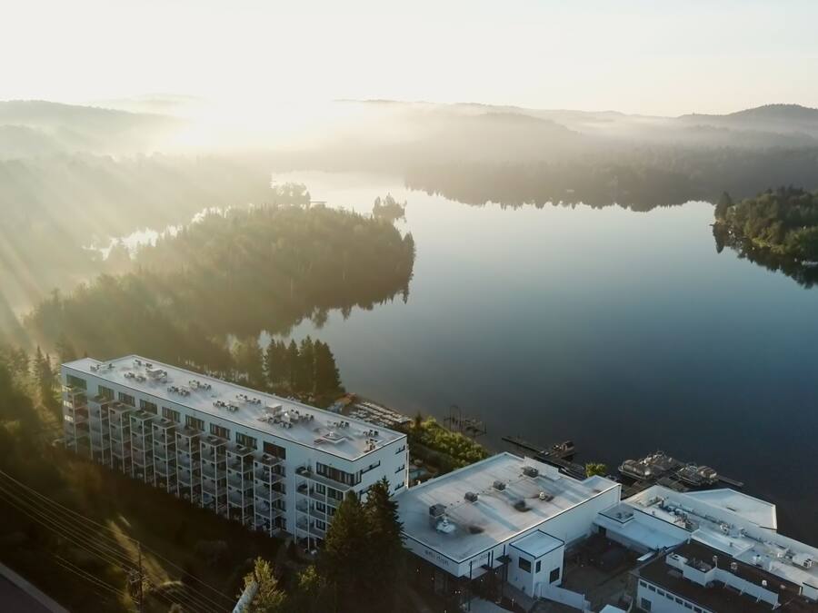 An aerial view of a modern hotel with multiple buildings, surrounded by a serene lake and forested hills.