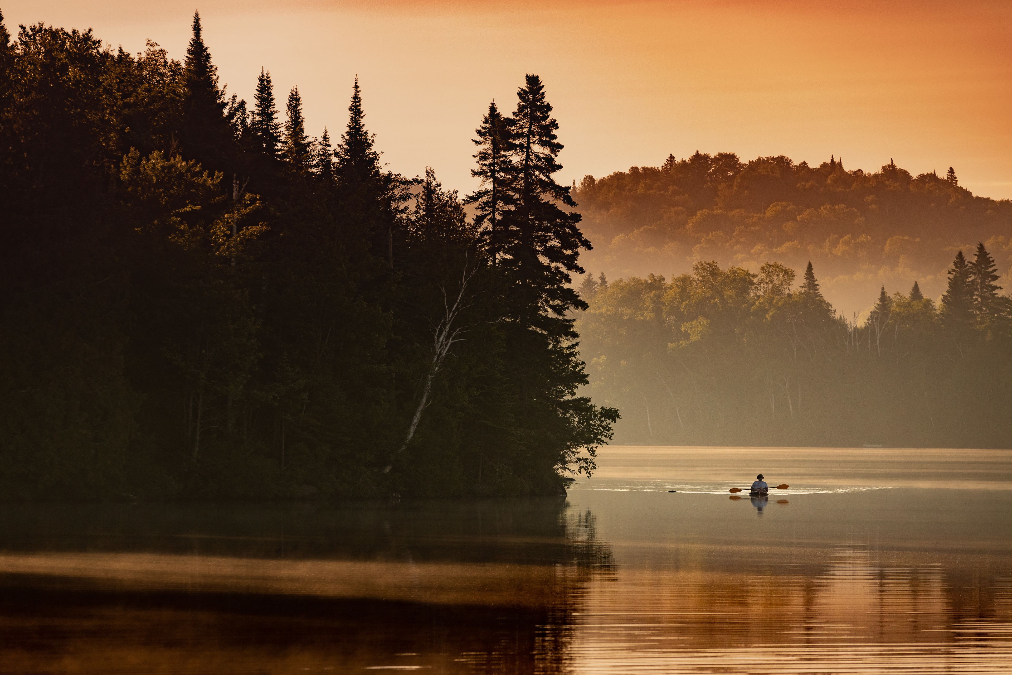Un lac paisible, avec un seul kayakiste aperçu au loin, entouré d’une forêt dense et enveloppé de brume.