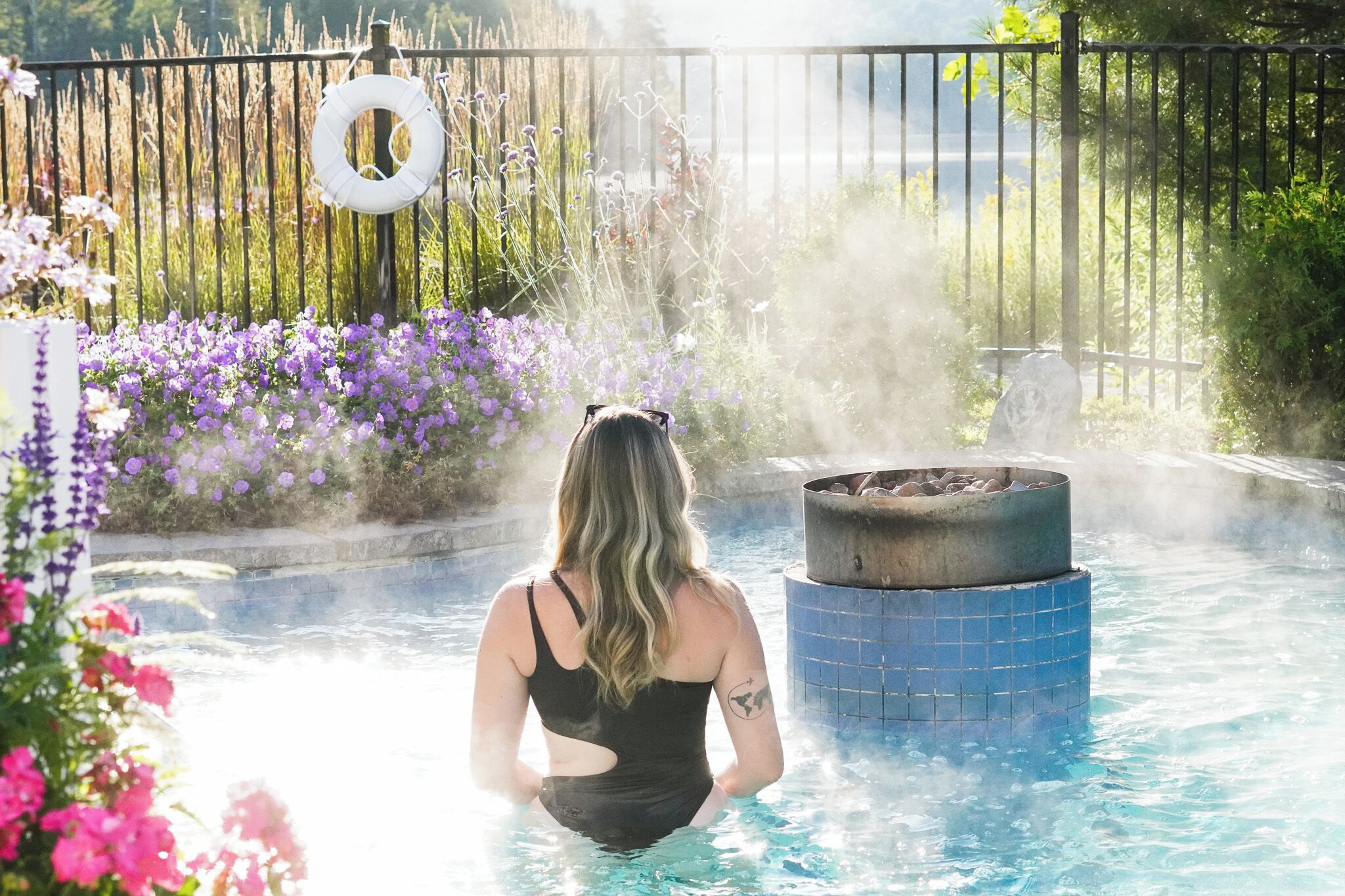 Una persona con traje de baño negro en una piscina con una fuente de fuego y una cerca con un columpio de neumático.