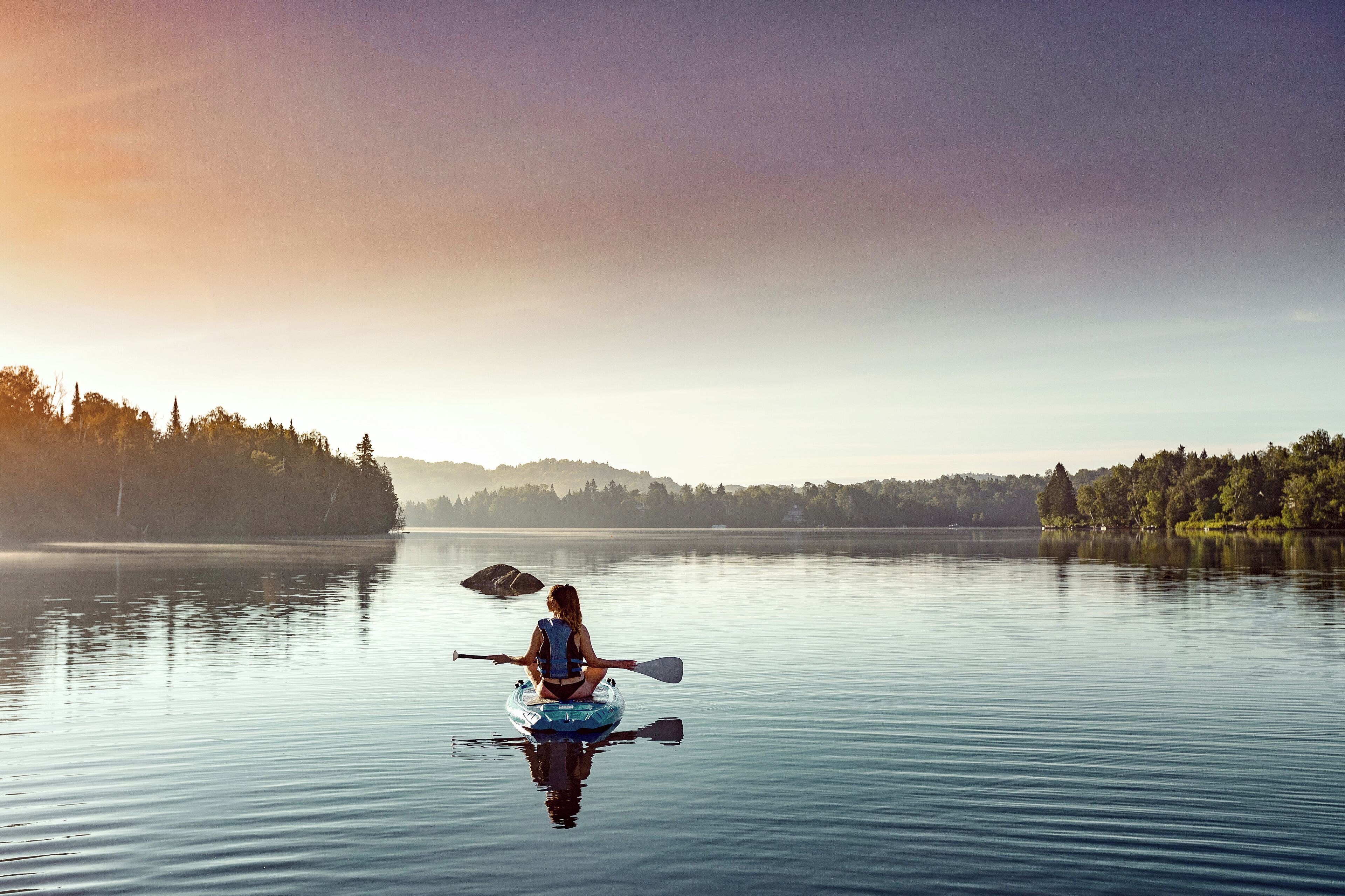 A person kayaking on a calm lake with a forested shoreline in the background.