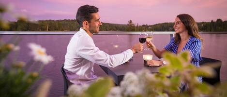 A couple toasting with wine glasses at a table with a view of a forested landscape.