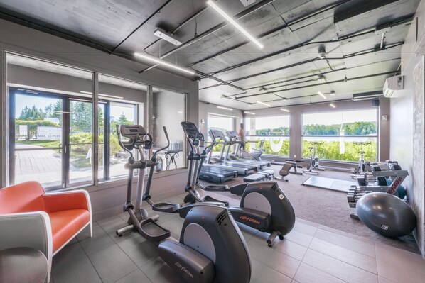 Un gimnasio con máquinas de cardio, bancos de pesas y un banco con vistas al exterior.
