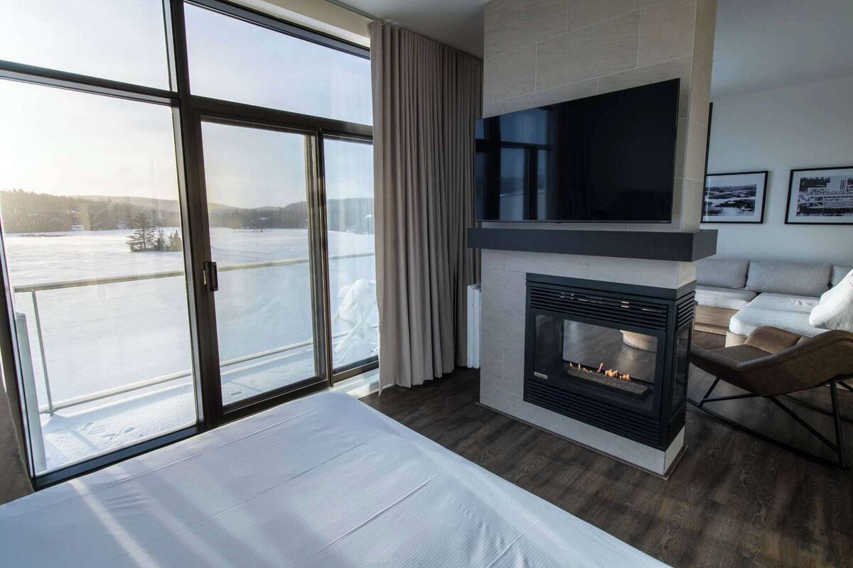 A modern hotel room with a large window, a fireplace, a flat-screen TV, and a bed.