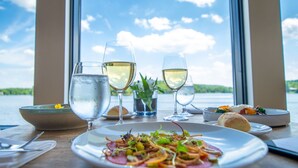 Ein Tisch mit drei Weingläsern, einer Salatschüssel und einem Teller Essen, mit Blick auf einen See durch große Fenster.