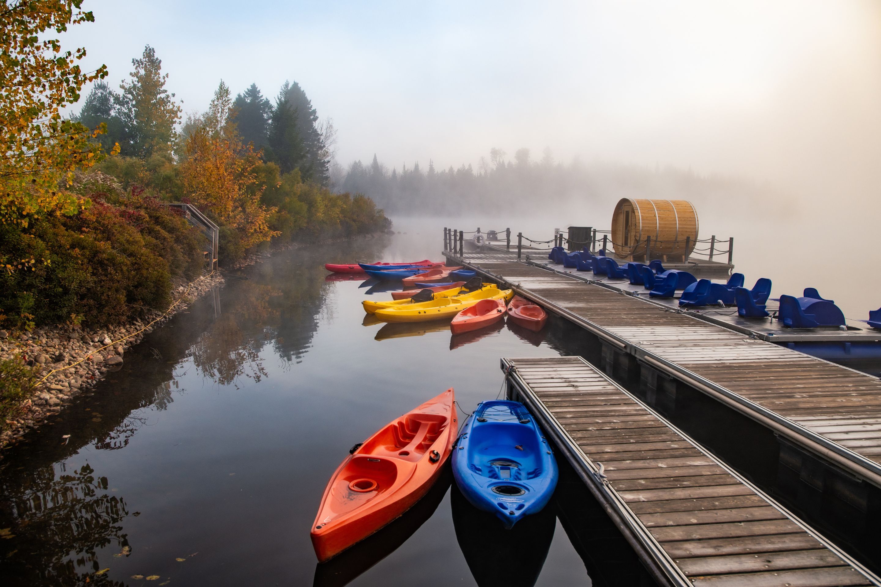 A calm river with kayaks docked on a wooden pier, surrounded by autumn foliage.