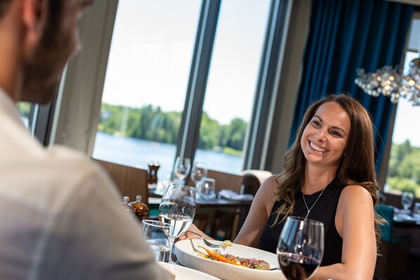 A dining table with a plate of food and wine glasses, a window with a view of a body of water, and a chandelier in the background.