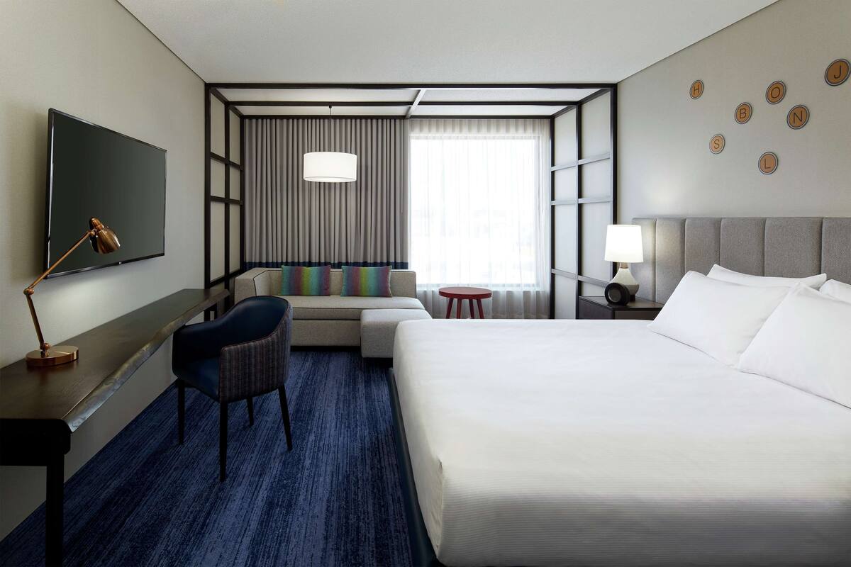 A hotel room with a large bed, a desk with a lamp, a chair, a sofa with colorful cushions, a small table, and a TV mounted on the wall.