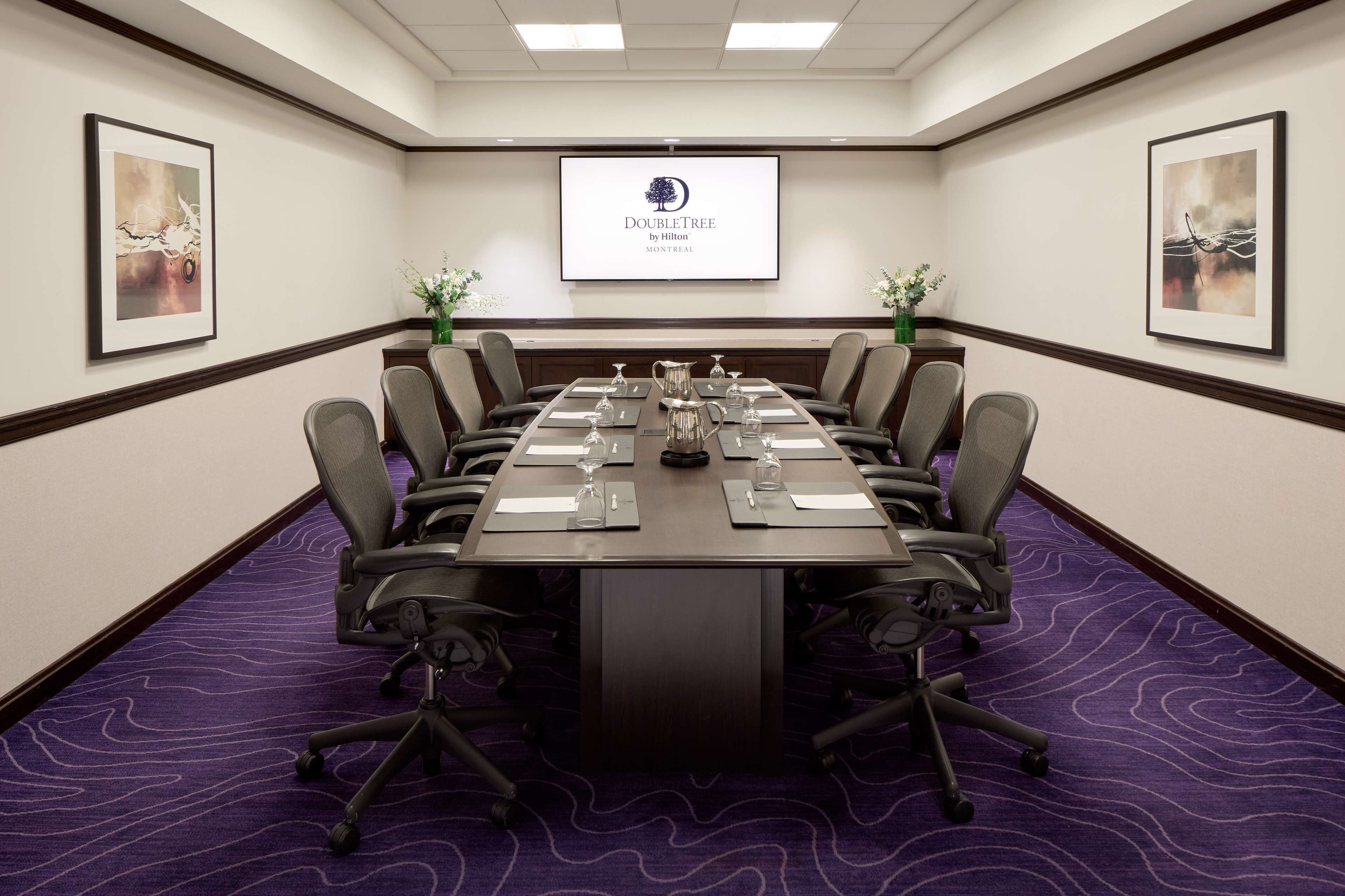 Egy tárgyalóterem, melyben egy hosszú asztal, székek és egy projektorvászon található, amelyen a DoubleTree by Hilton logója látható.