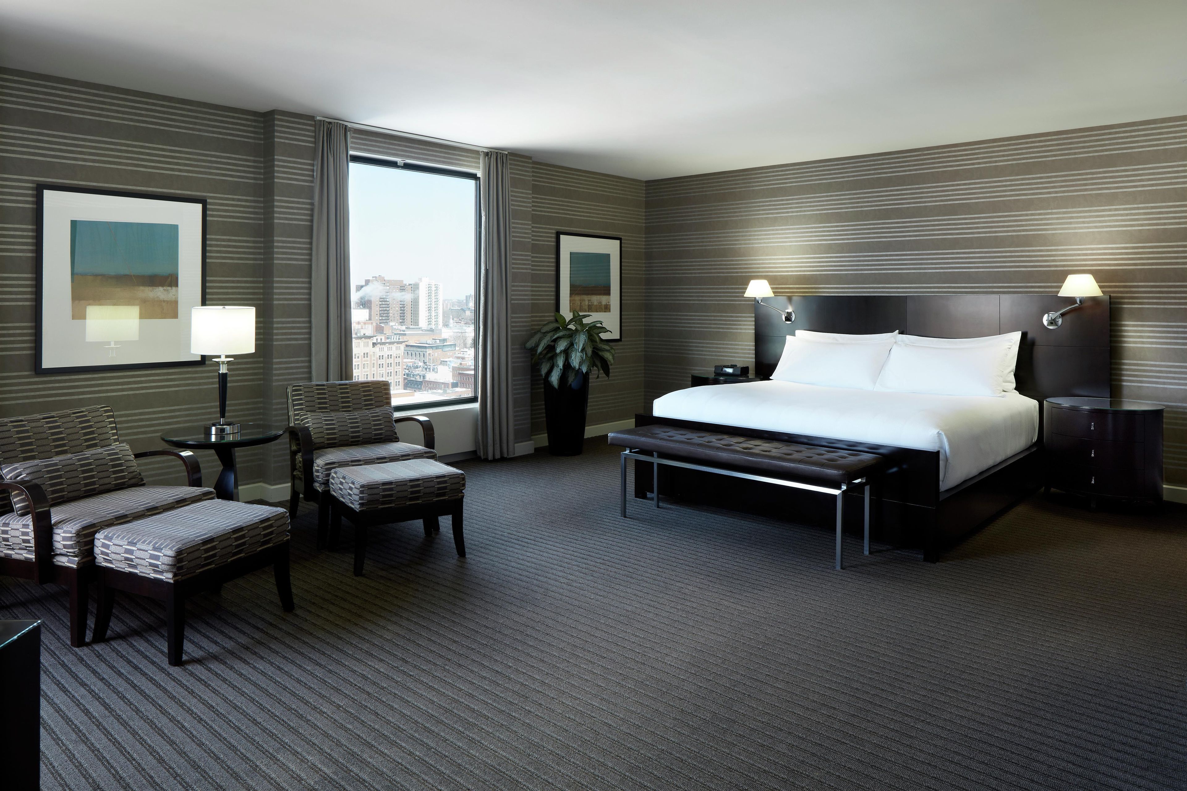 A hotel room with a large bed, two armchairs, a small table, a potted plant, and a view of the city through the window.