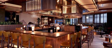 Un espace bar moderne doté d’un long comptoir, de nombreux tabourets et d’un plafond haut avec des poutres apparentes.