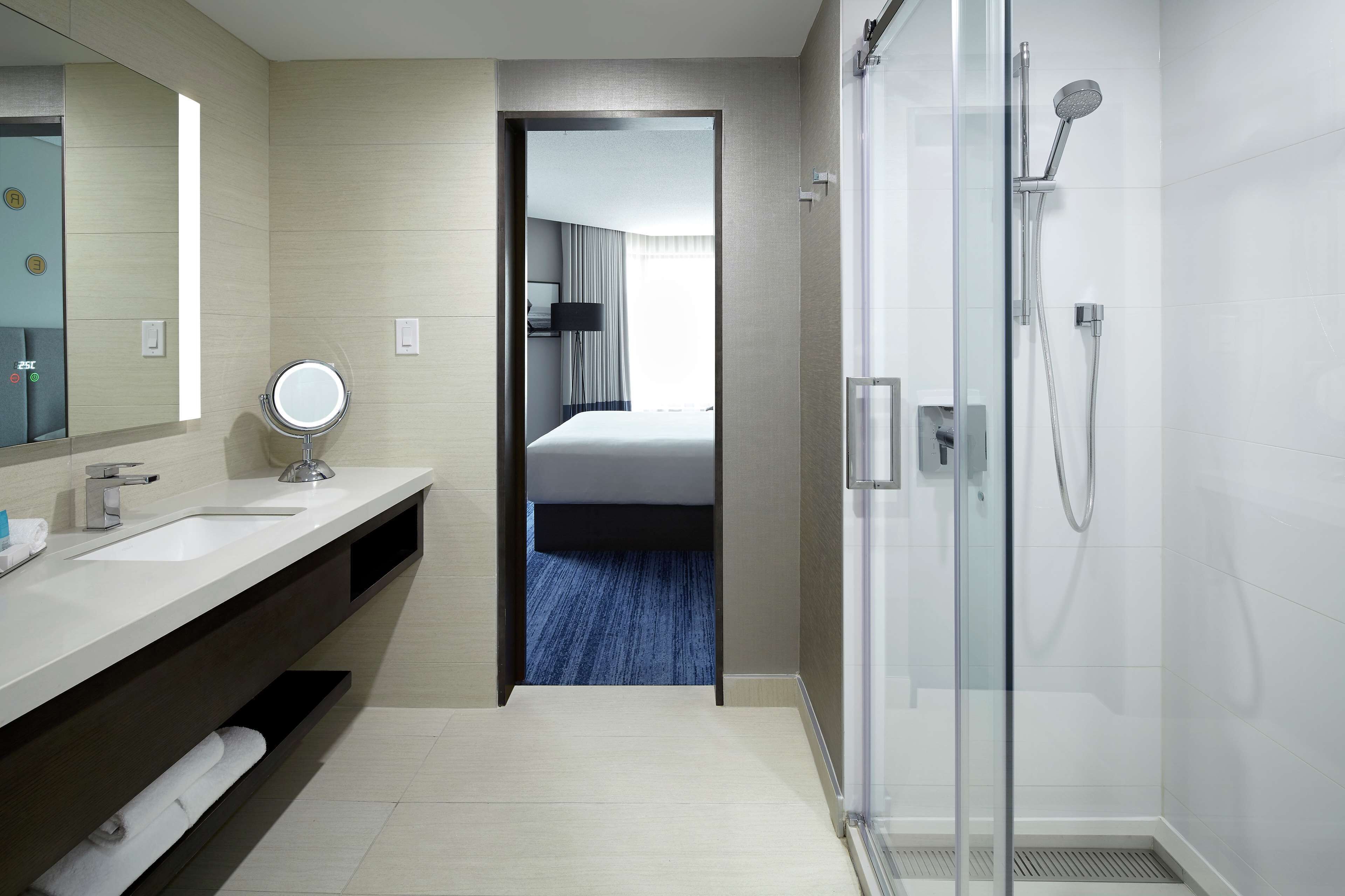 Une salle de bain moderne dans un hôtel, dotée d’une douche en verre, d’un grand miroir et d’une vue sur la chambre à coucher, visible à travers une porte ouverte.
