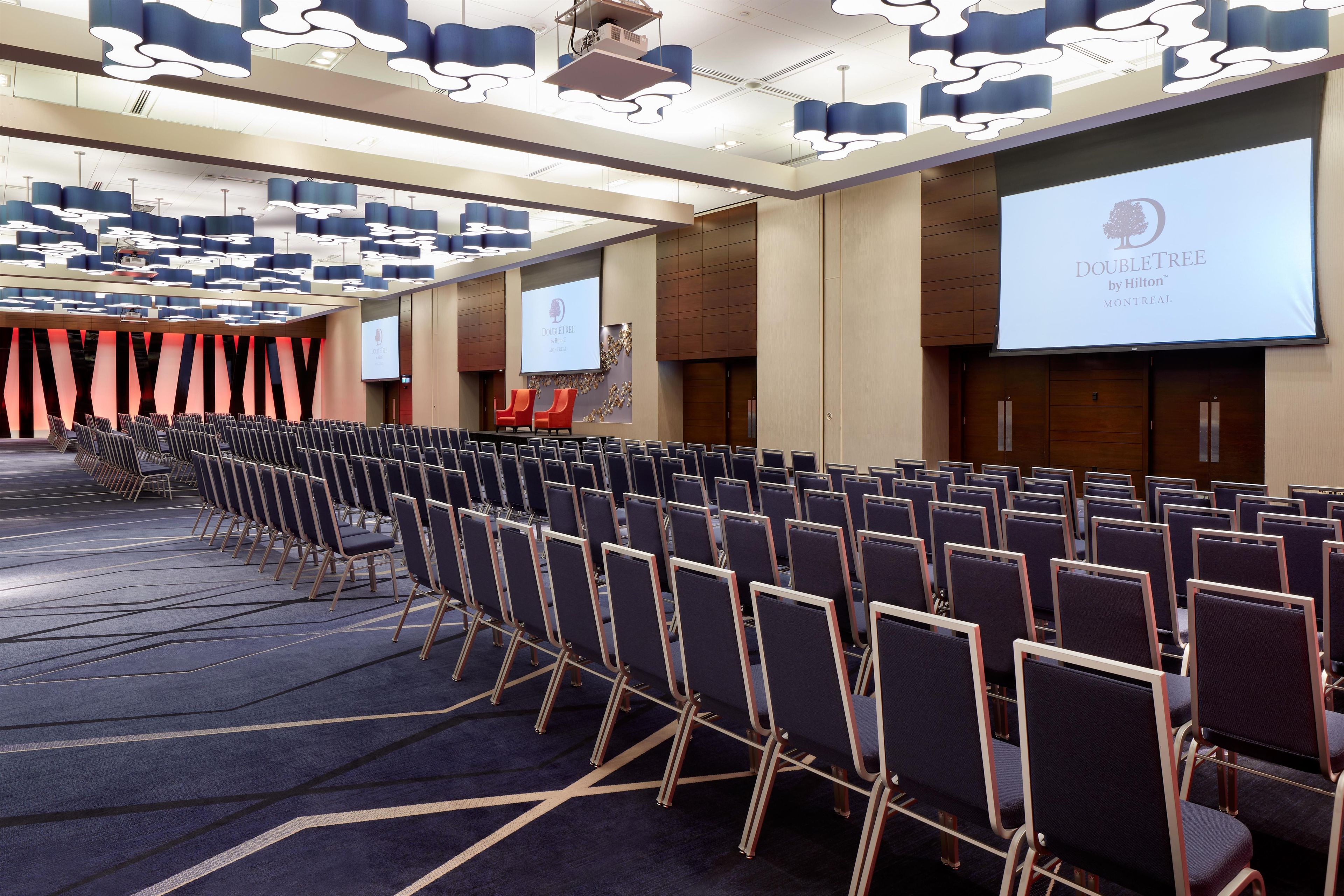 Konferenssitila, jossa on riveissä tuoleja, suuri näyttö, jossa näkyy DoubleTree by Hilton -hotelliketjun logo, ja moderni kattorakenne.