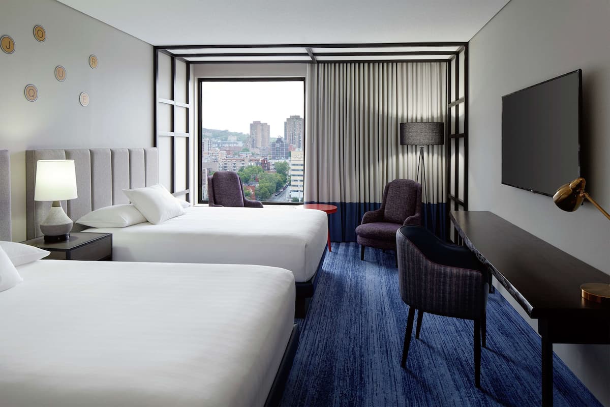 A hotel room with two beds, a desk, a chair, a TV, and a view of the city.