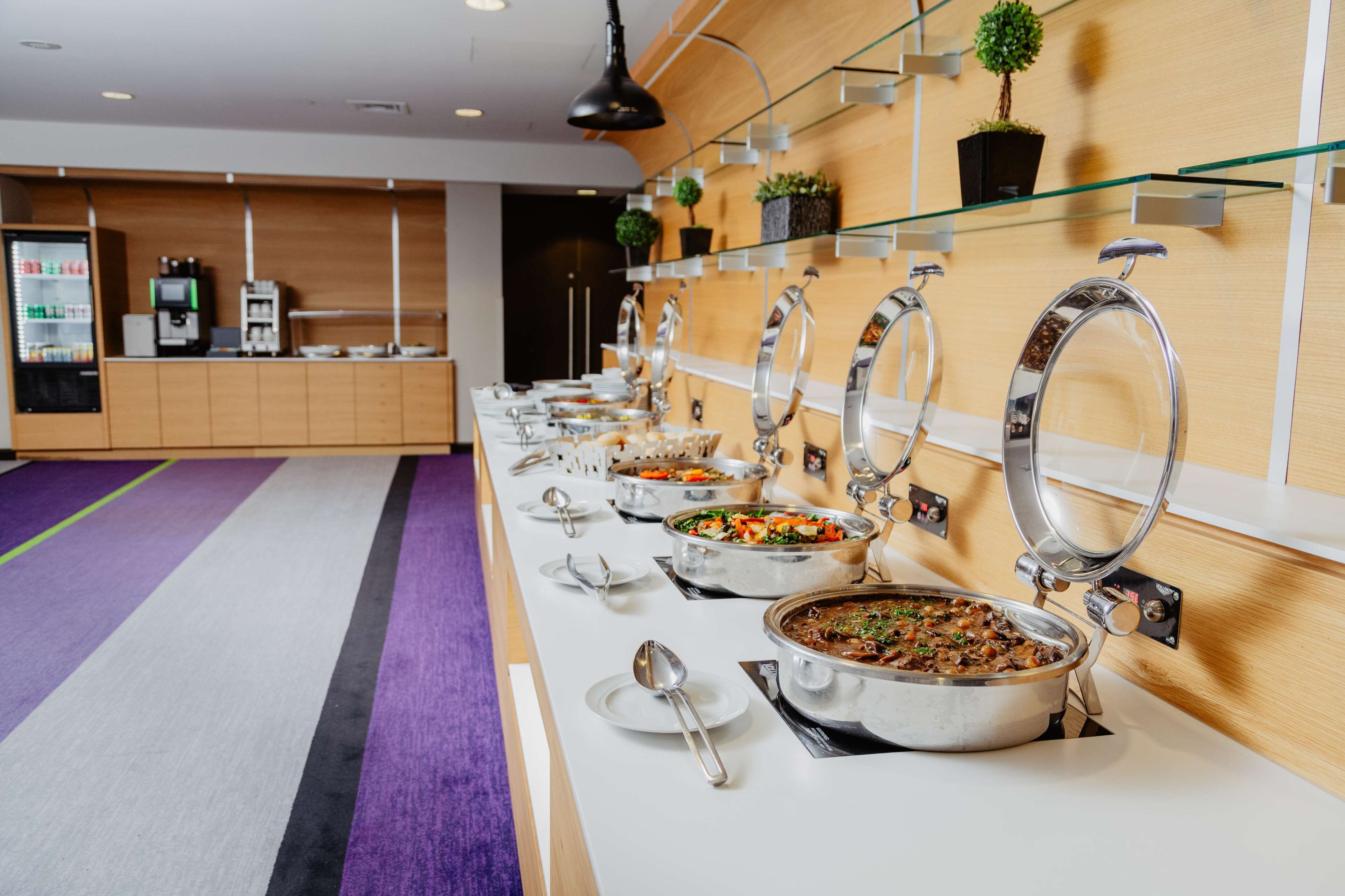Een buffetopstelling met meerdere serveerpunten, een koffieapparaat en een zithoek met een paars vloerkleed.