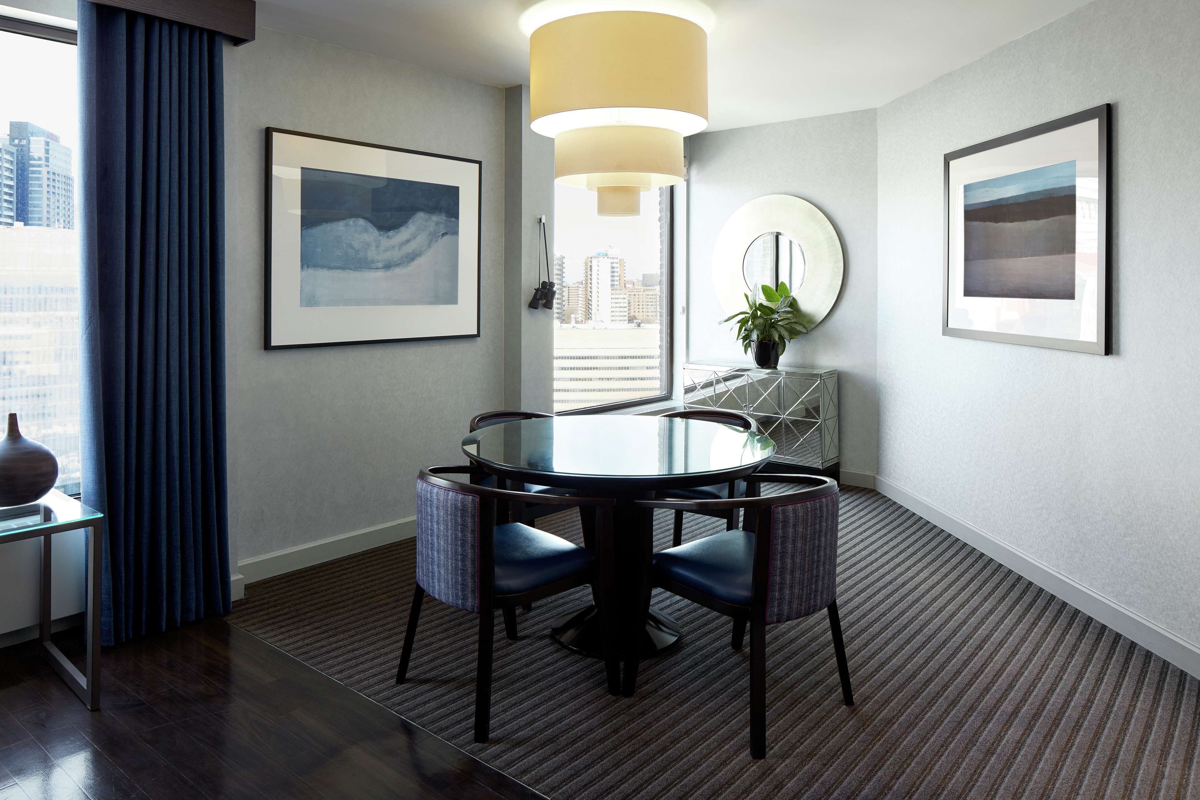 A modern hotel room with a round dining table, blue chairs, a large window with curtains, and framed artwork on the walls.