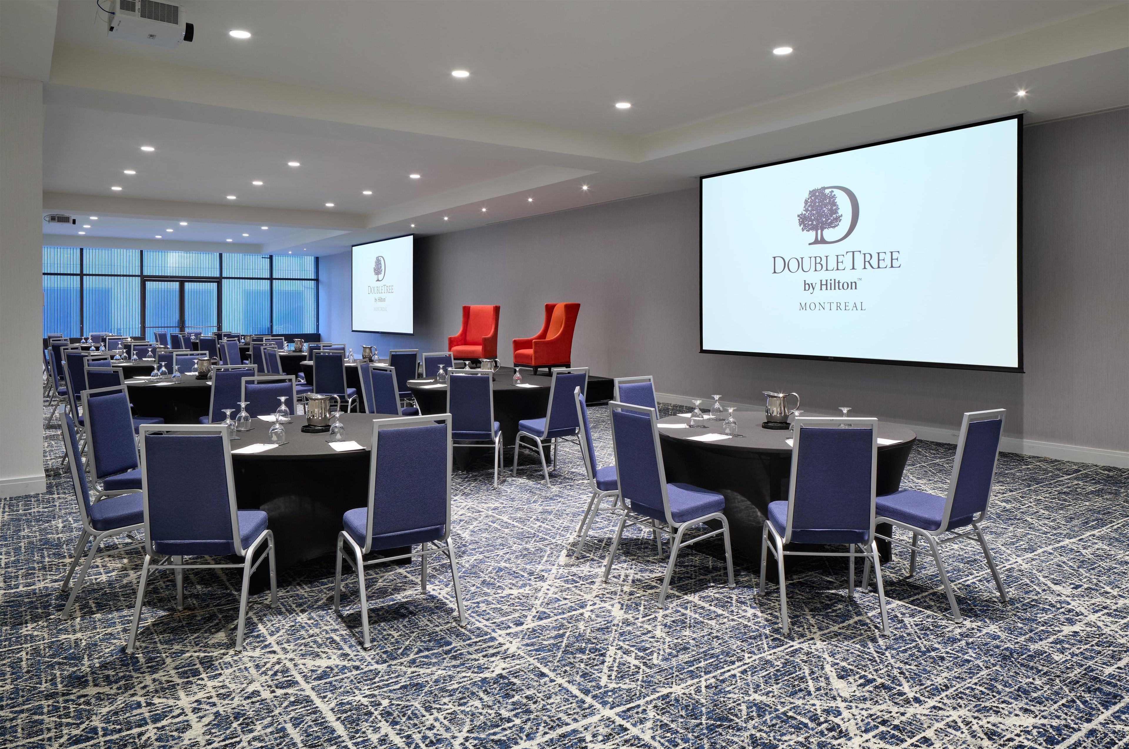 Une salle de conférence dotée de plusieurs tables et chaises, d’un grand écran affichant le logo de la chaîne DoubleTree by Hilton, et d’un sol recouvert de moquette.