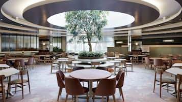 Un vaste espace intérieur aménagé avec différents types de sièges, notamment des tables et des chaises rondes, et un arbre central.