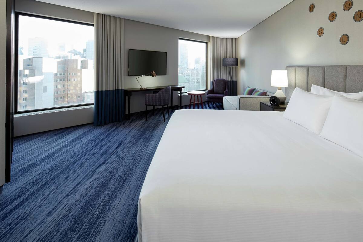 A hotel room with a large bed, a desk with a chair, a TV, and a view of the cityscape through large windows.