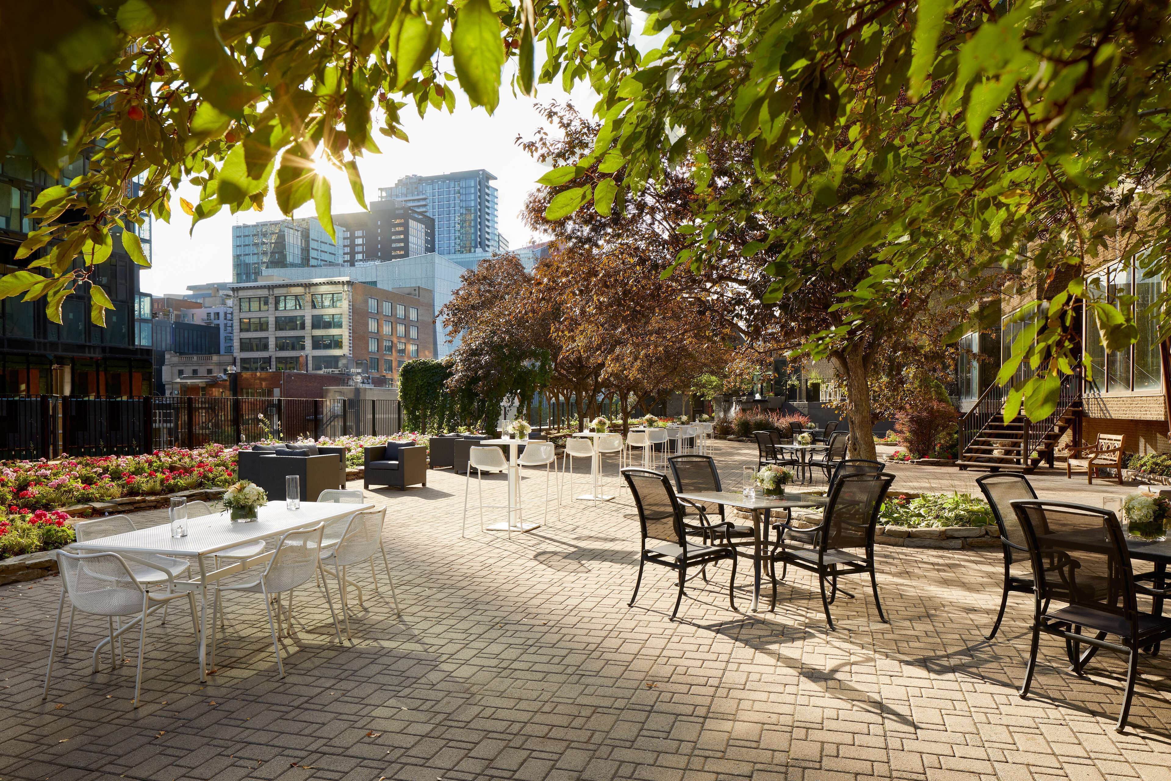 An outdoor patio area with tables and chairs, surrounded by plants and trees, with a cityscape in the background.