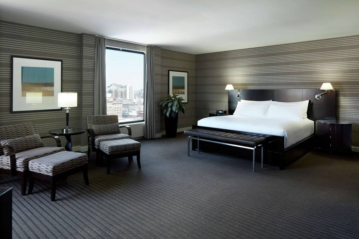 A hotel room with a large bed, two chairs, a small table, and a view of the city.