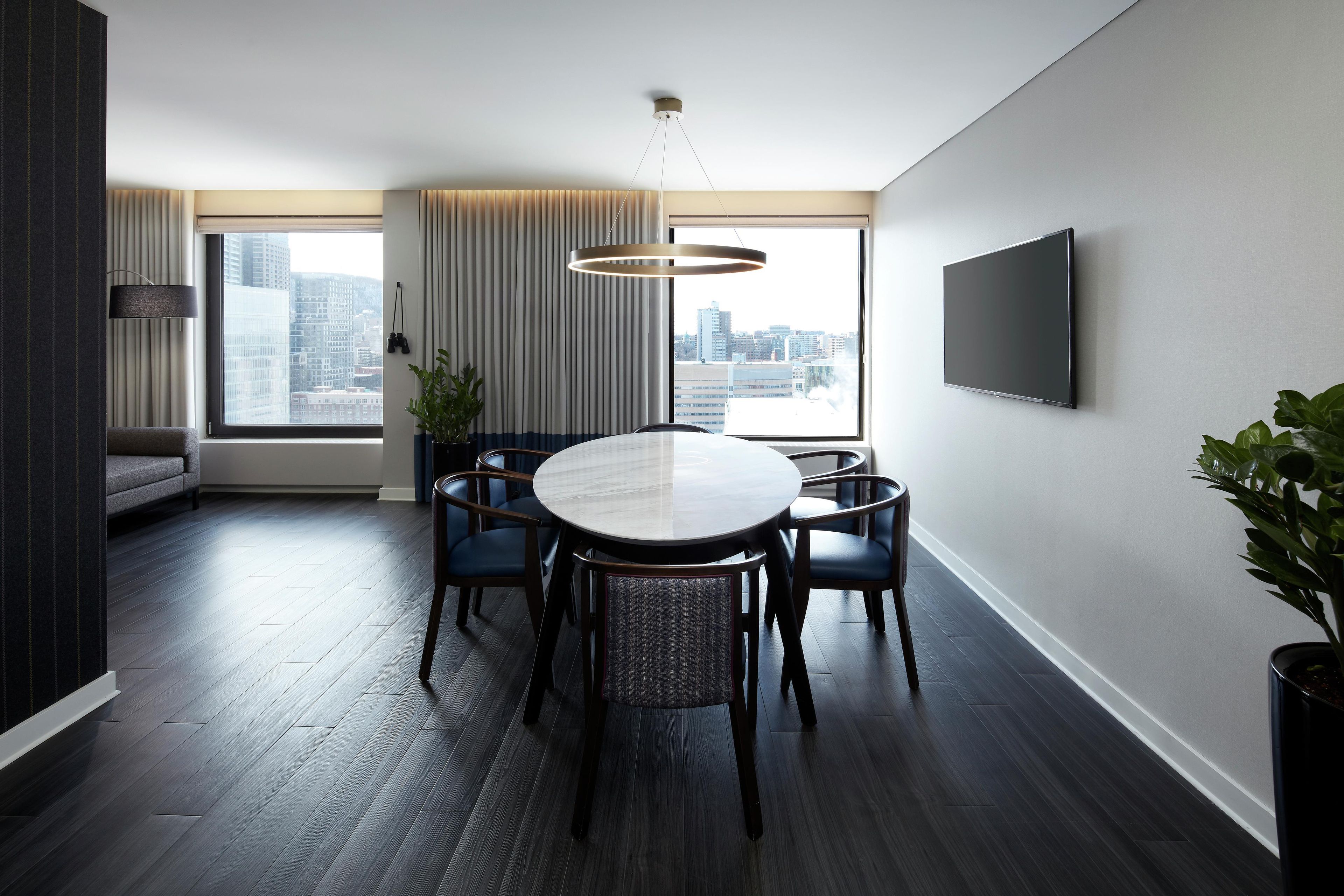 A modern dining area with a round table, dark wood flooring, and a city view through large windows.