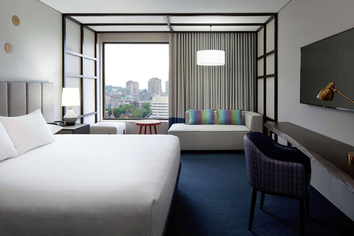 A hotel room with a large bed, a sofa, a desk, and a chair. There is a TV mounted on the wall, a lamp, and a view of the city through the window.