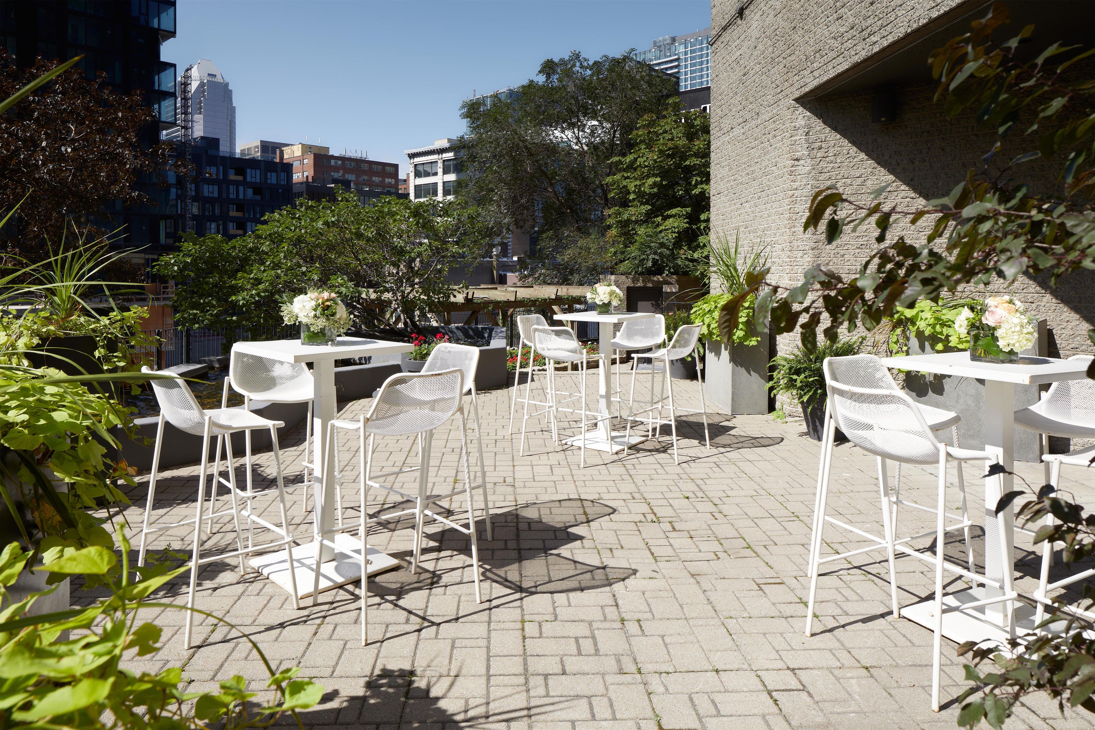 An outdoor patio with white metal chairs and tables, surrounded by greenery and a cityscape in the background.