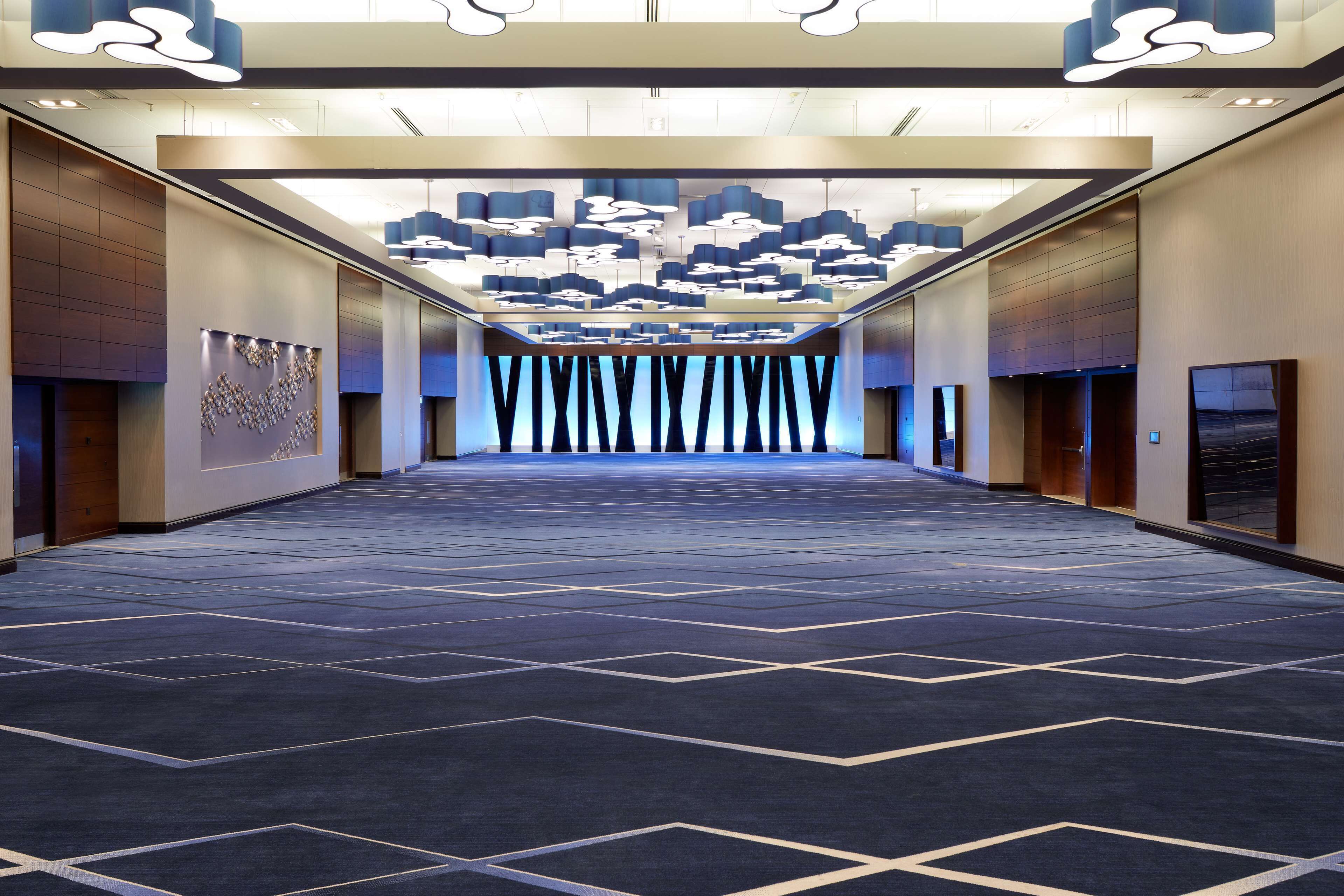 Un vaste couloir d’hôtel agrémenté d’un tapis à motifs, de luminaires modernes et d’un plafond au design géométrique.