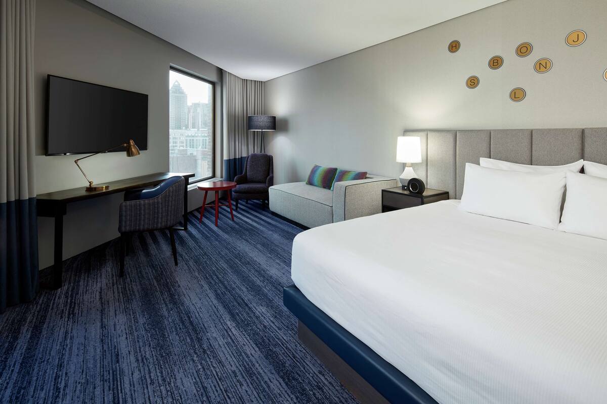 A hotel room with a large bed, a desk with a chair, a sofa, a TV, and a city view.