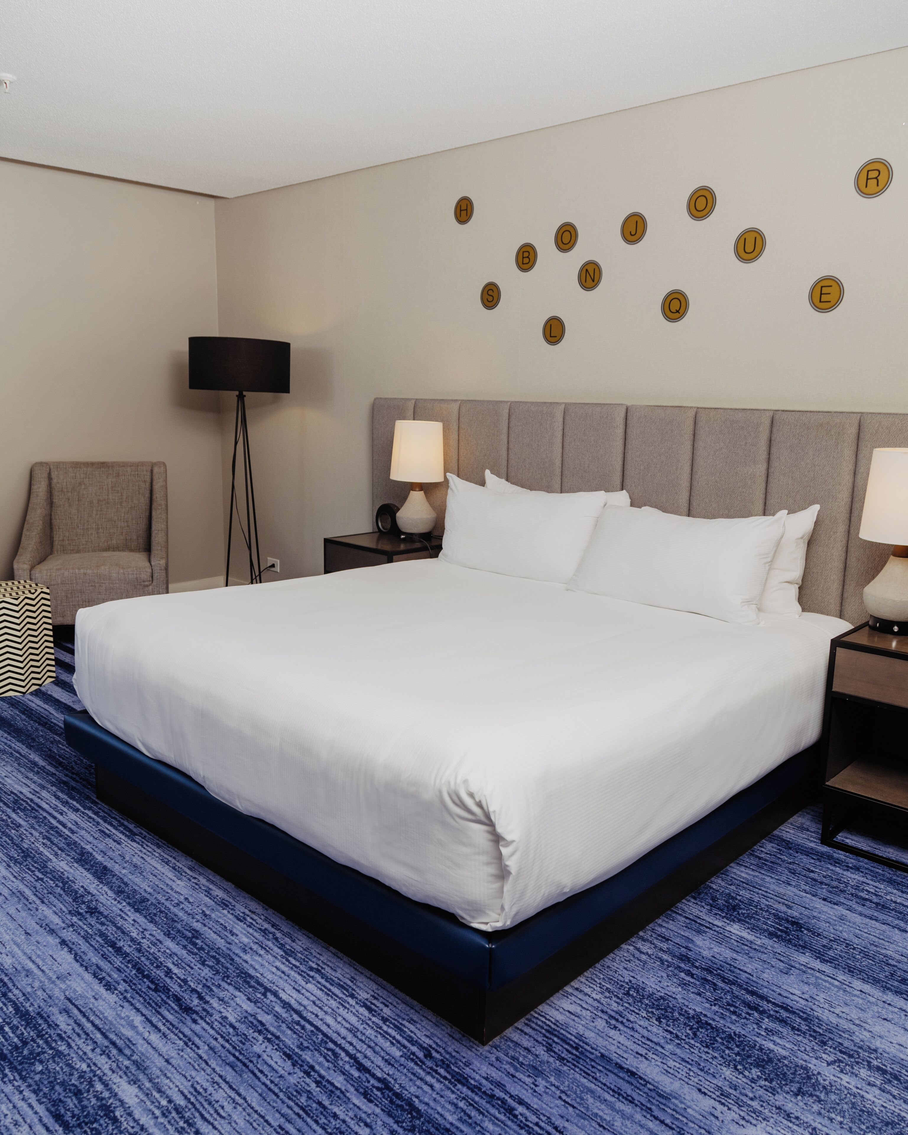 Une chambre d’hôtel avec un grand lit, une chaise, une lampe sur pied, une table de chevet avec une lampe, et un mur décoré de motifs circulaires.