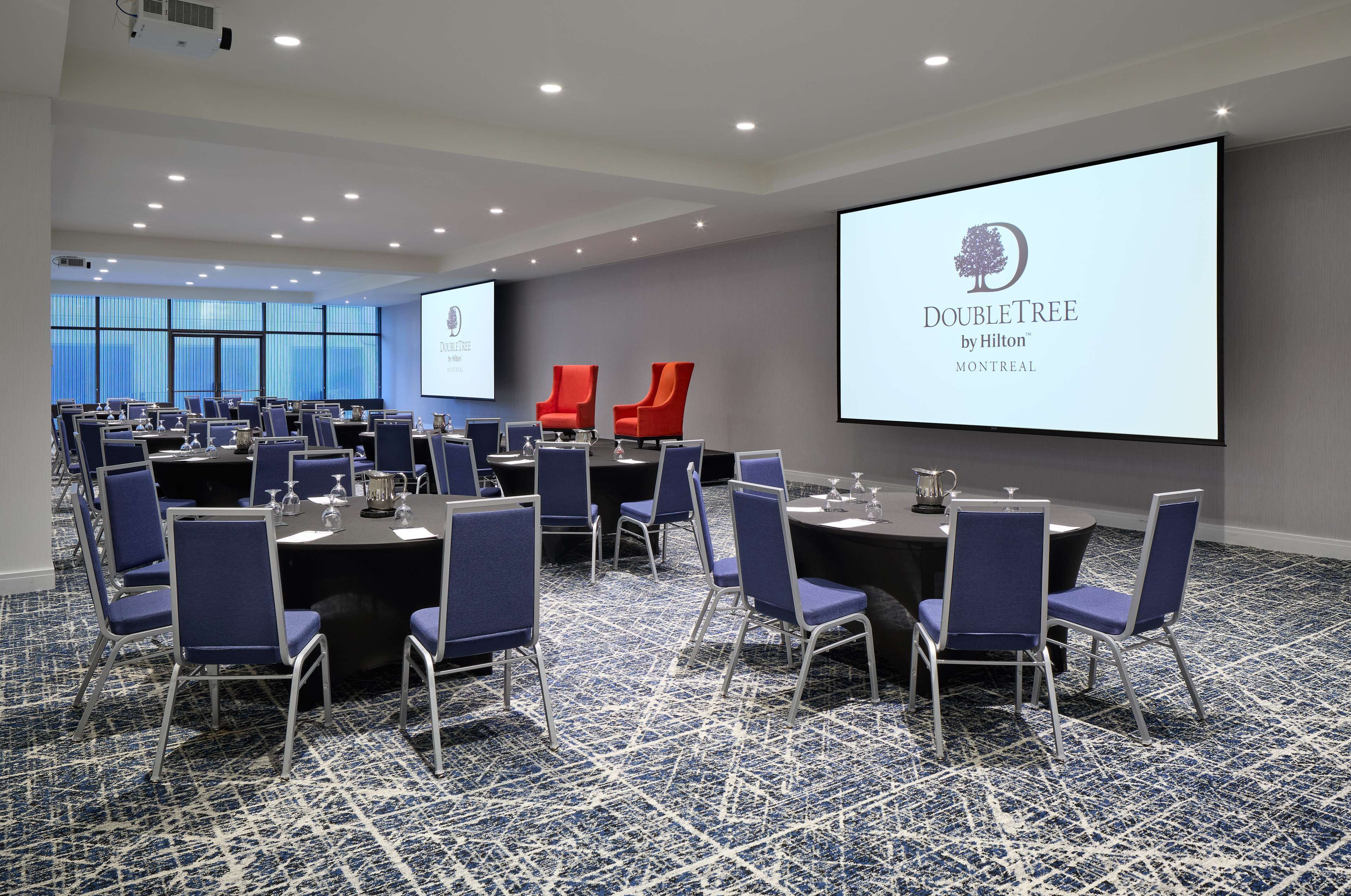 A conference room with multiple tables and chairs, a large screen displaying the DoubleTree by Hilton logo, and a carpeted floor.
