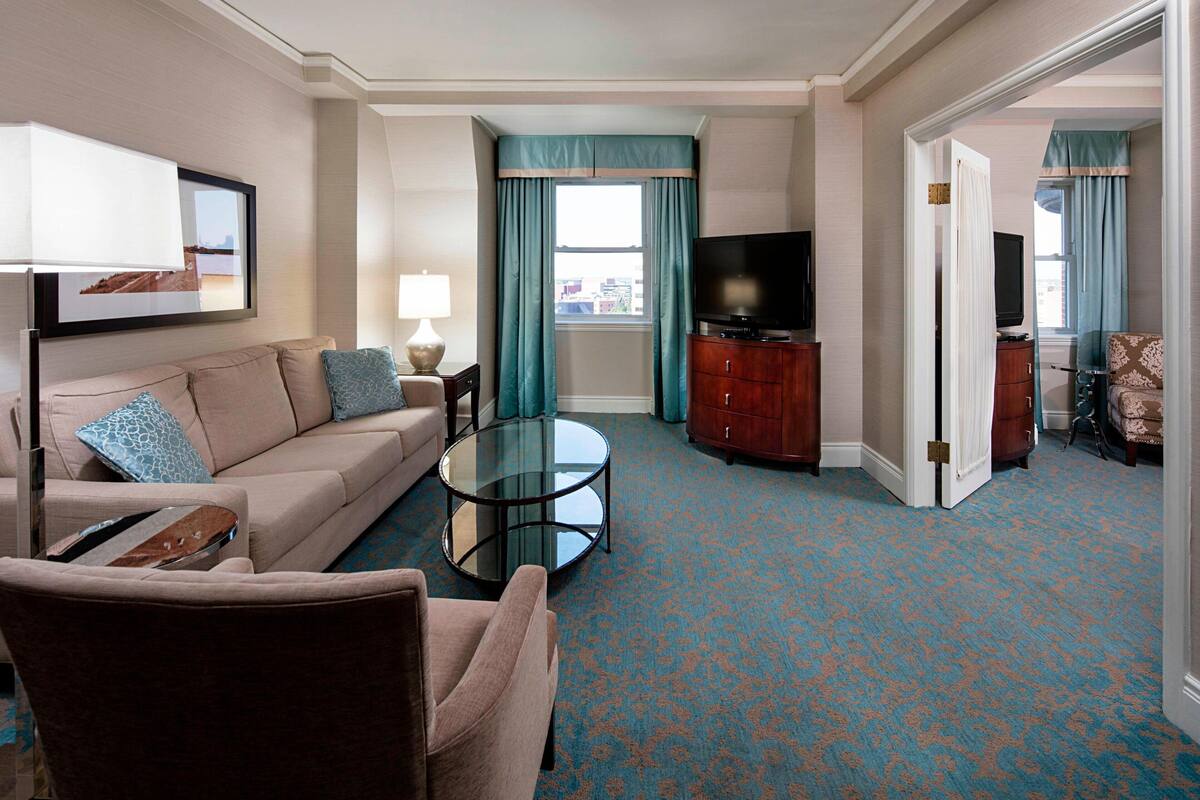 Quarto de hotel com sofá, poltrona, mesa de centro de vidro e televisão sobre um armário de madeira.