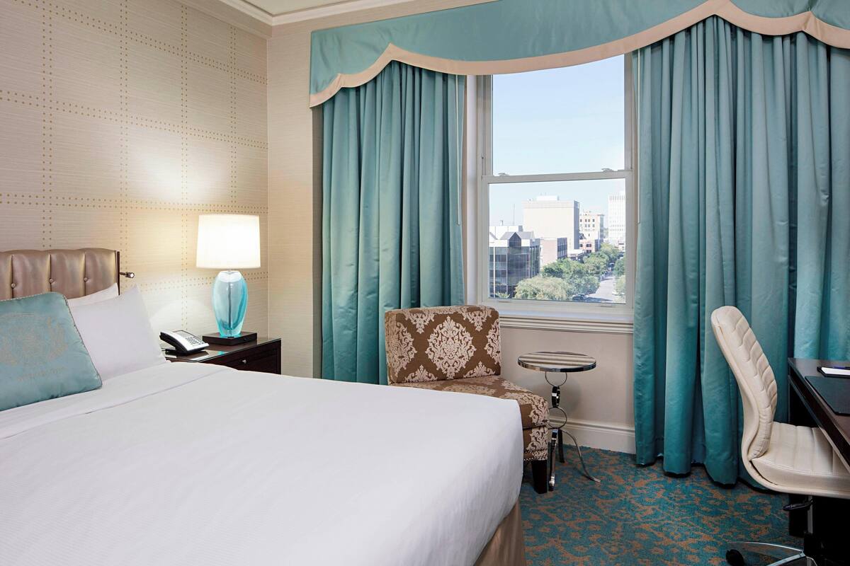 Quarto de hotel com uma cama grande, uma escrivaninha, uma cadeira, uma mesinha e vista da cidade através de uma janela com cortinas azul-turquesa.