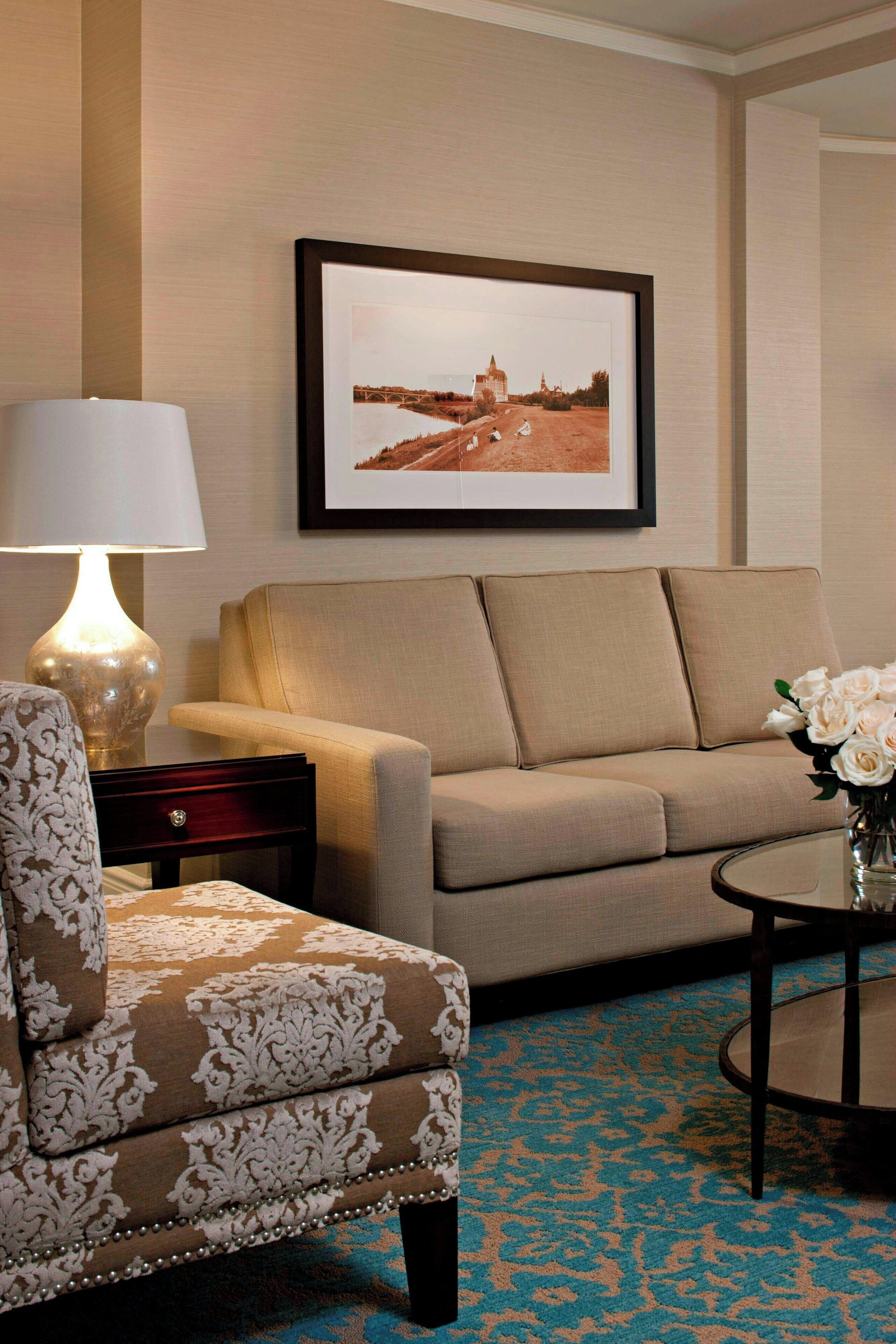 A hotel room with a beige sofa, a patterned armchair, a small table with a vase of flowers, a lamp, and a framed picture on the wall.