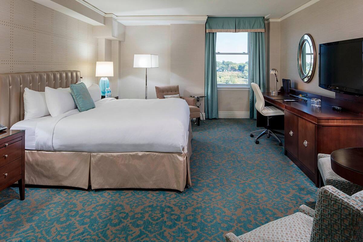 Quarto de hotel com uma cama grande, uma escrivaninha com televisão, uma cadeira e abajur.