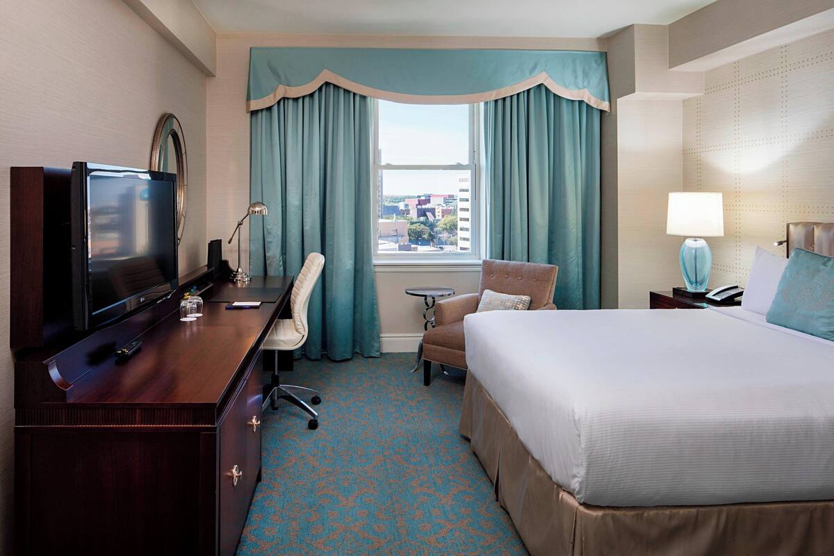 Quarto de hotel com cama, escrivaninha com televisão, cadeira, mesa pequena e janela com cortinas.