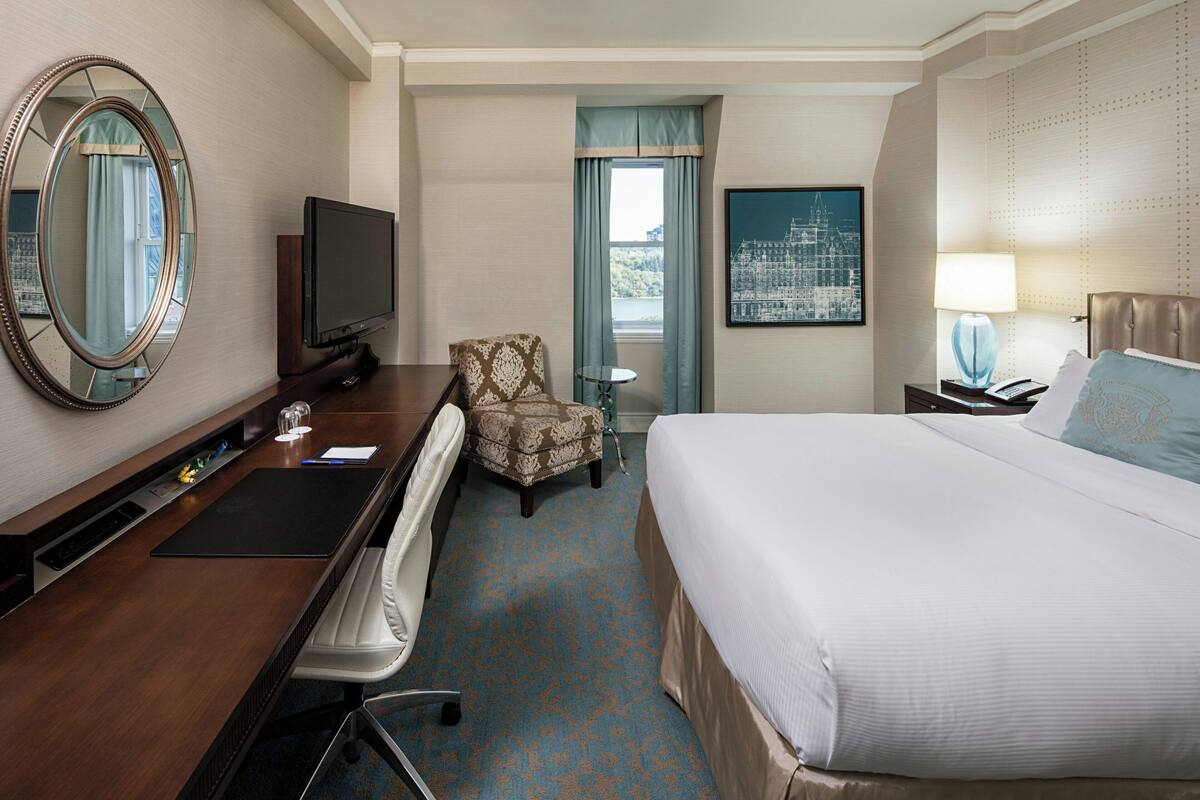 Quarto de hotel com uma cama grande, uma escrivaninha com cadeira, televisão, mesa pequena e janela com cortinas.