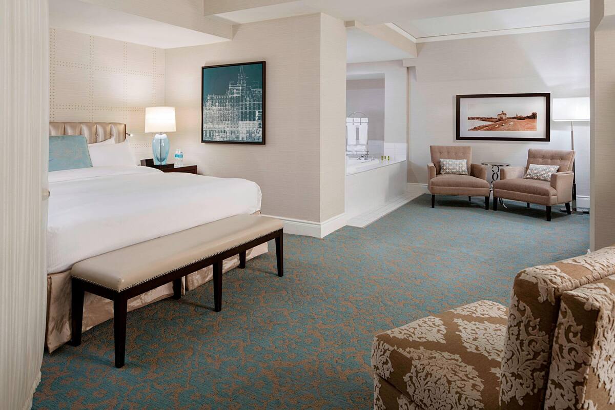 Quarto de hotel com uma cama grande, um banco, um sofá e duas poltronas. Há quadros nas paredes e uma mesinha com um abajur.