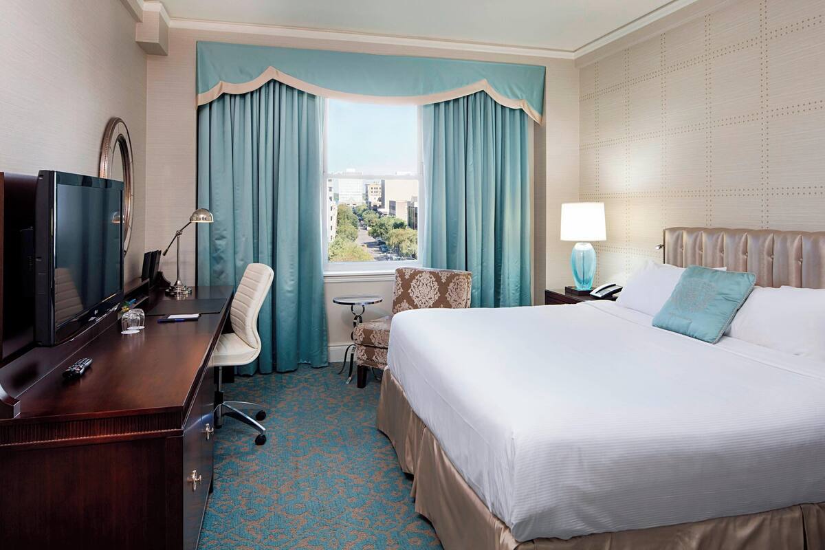 Quarto de hotel com uma cama grande, uma escrivaninha com televisão, uma cadeira, uma mesinha e uma janela com cortinas.