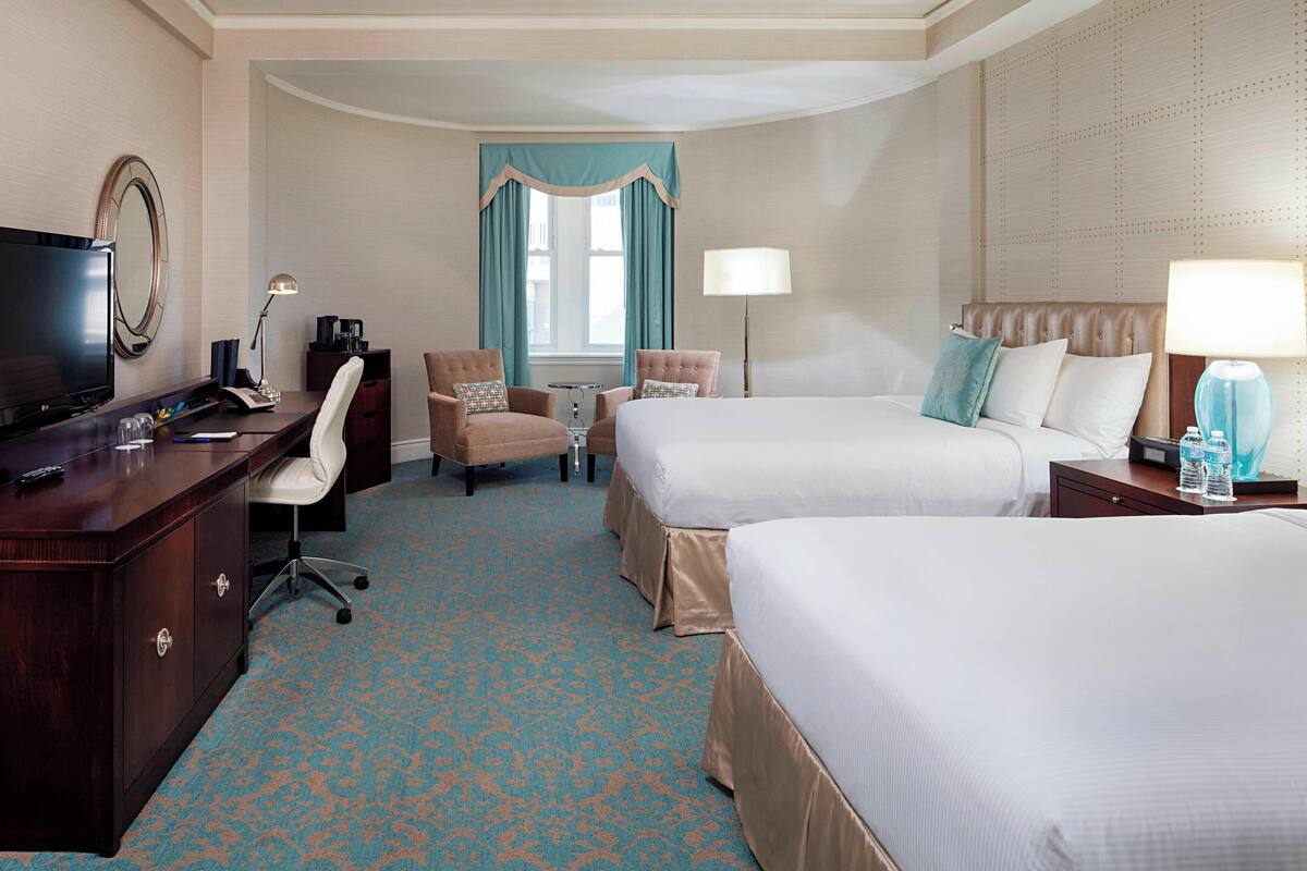 Quarto de hotel com duas camas, uma escrivaninha com televisão, uma cadeira, um abajur e uma janela com cortinas.