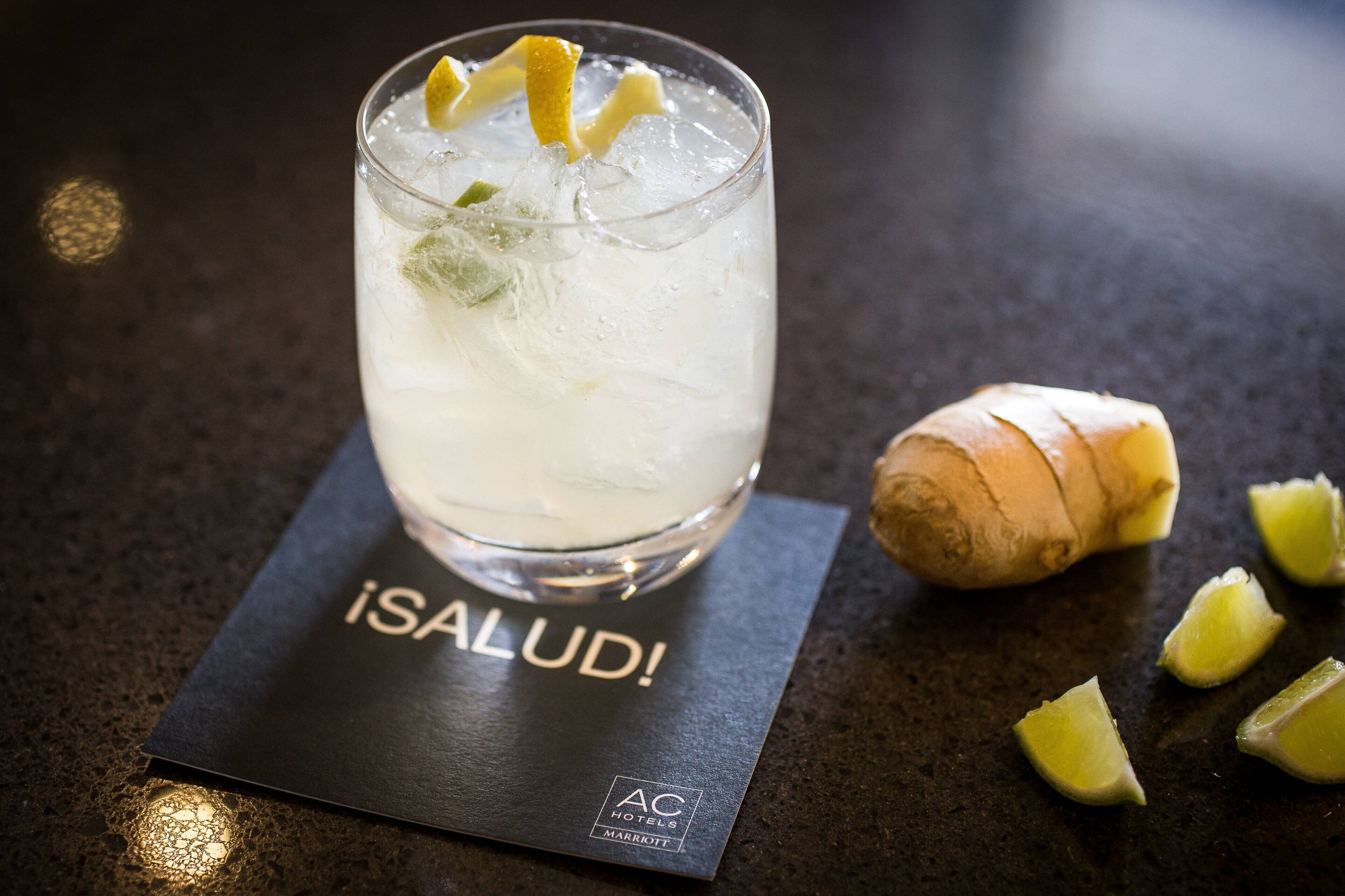A glass of iced beverage with lime slices and mint, placed on a black coaster with "iSALUD!" and "AC HOTELS MARROTT" text. A ginger root and lime wedges are nearby.
