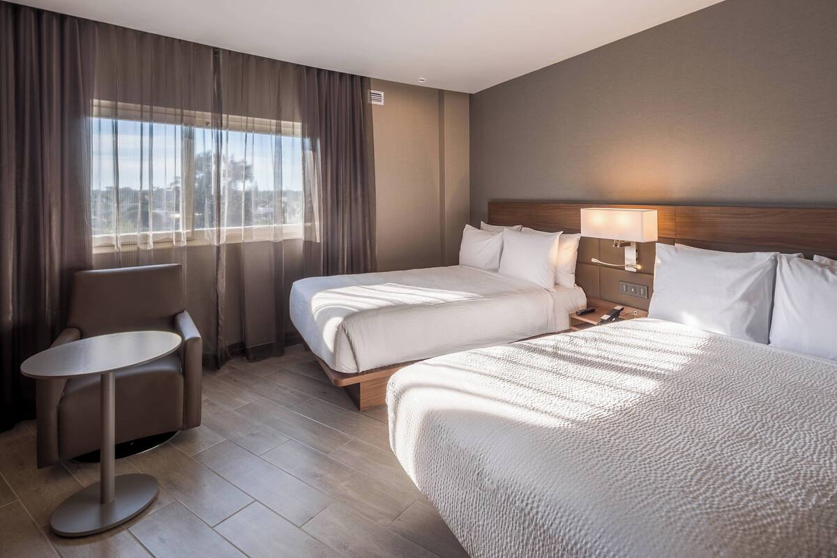 Une chambre d’hôtel avec deux lits, une petite table ronde et une vue sur la ville grâce à de grandes fenêtres.