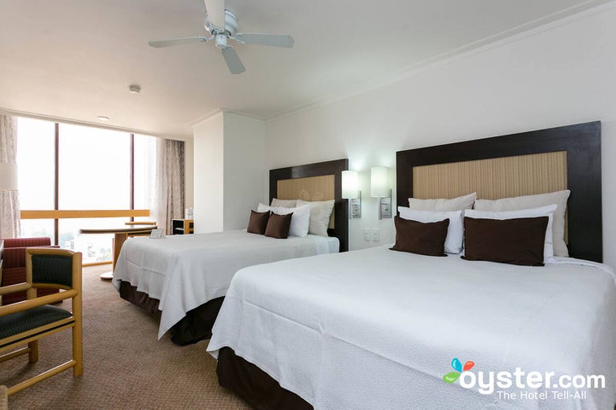Quarto de hotel com duas camas, uma escrivaninha, uma cadeira e um ventilador de teto.