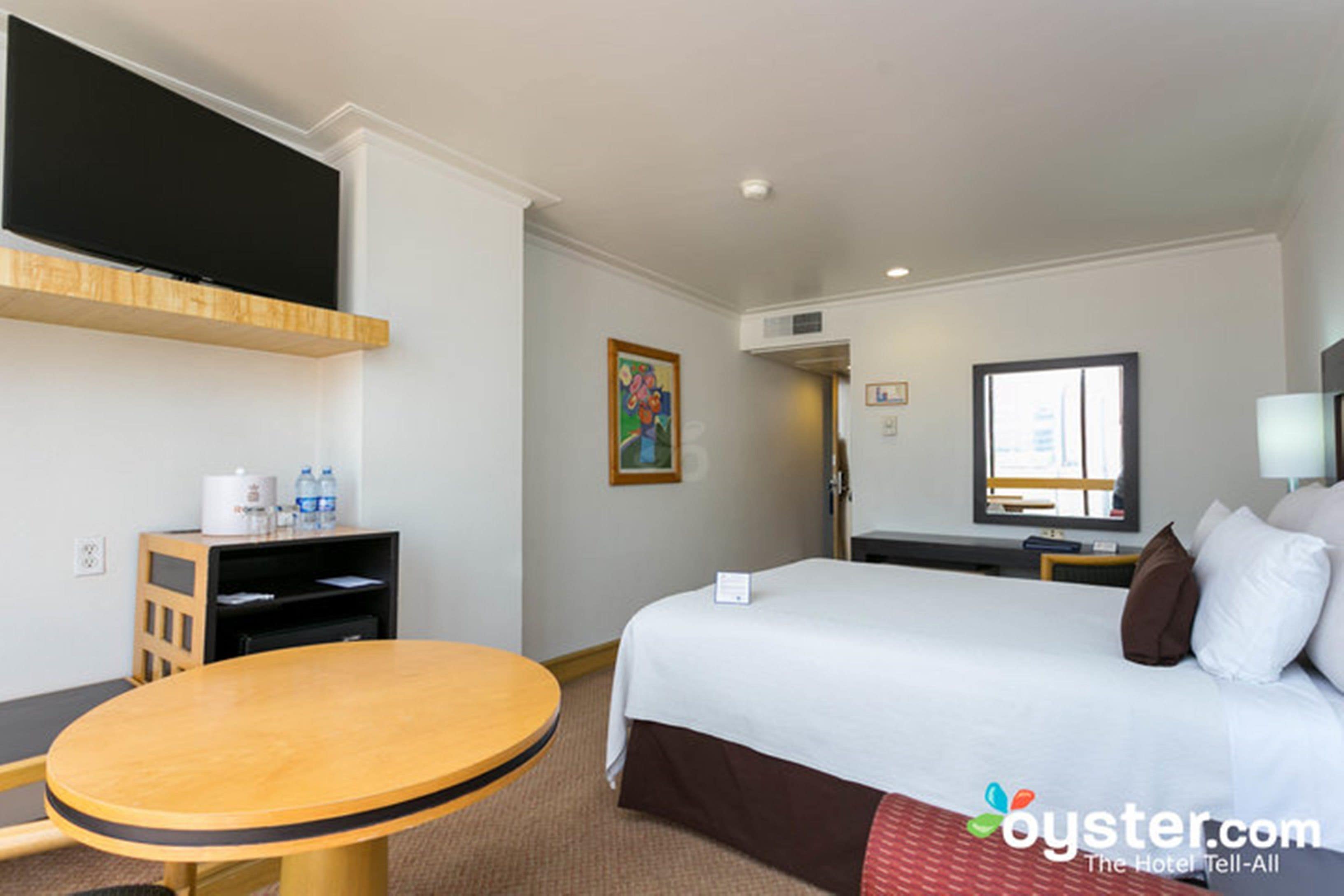 A hotel room with a bed, a TV mounted on the wall, a small table, and a window.