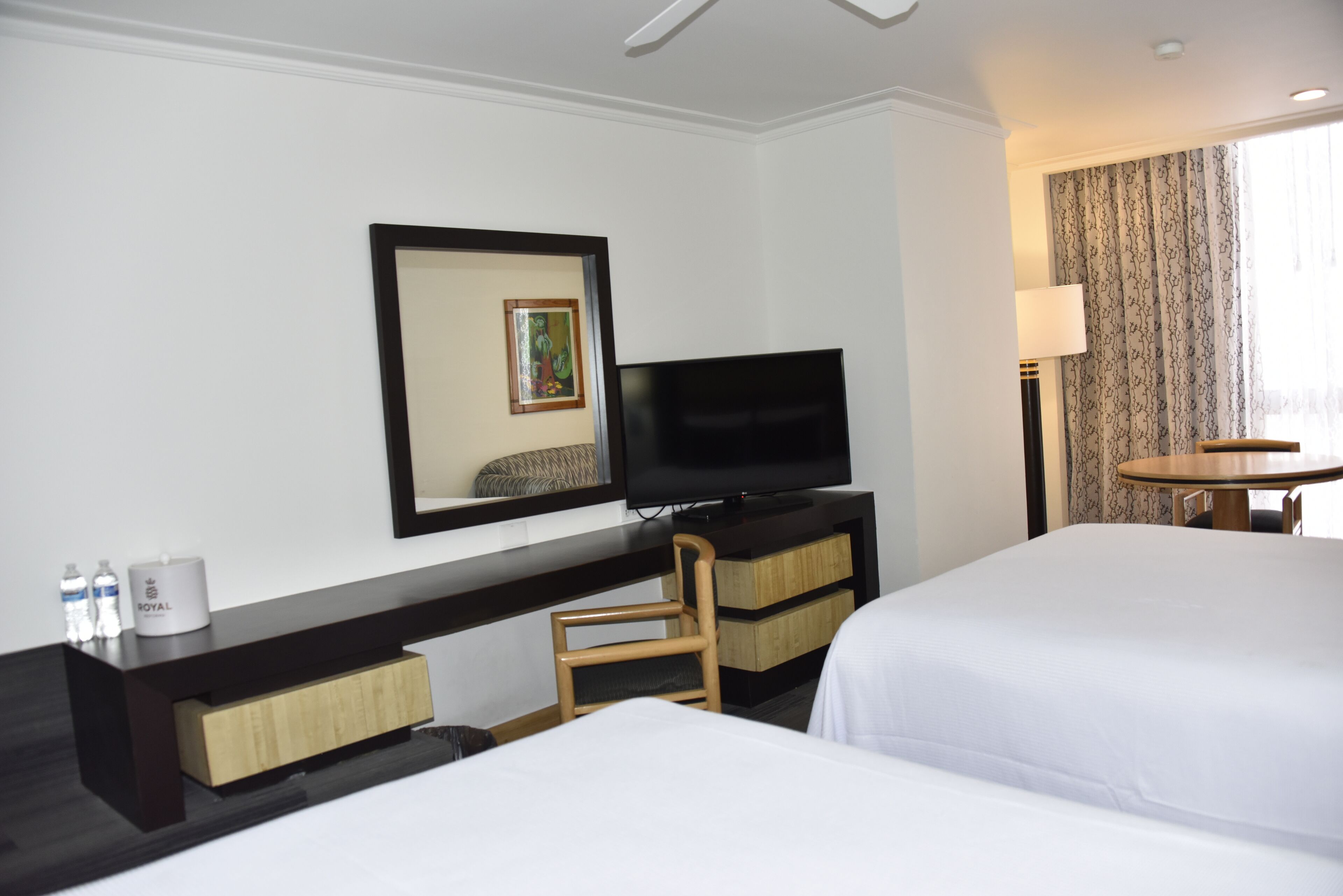 A hotel room with two beds, a TV, a desk with a chair, a lamp, and a window with curtains.