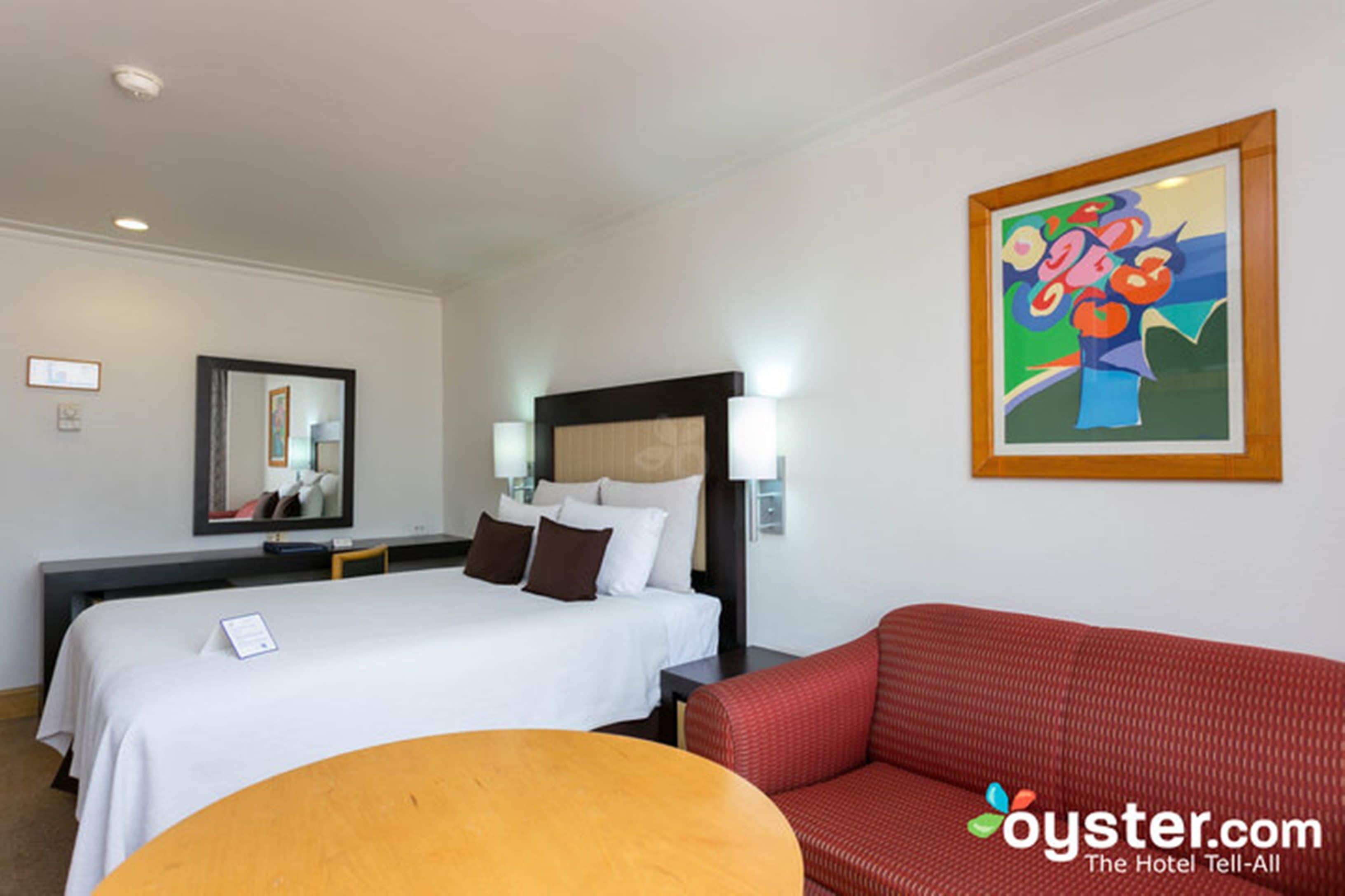 Une chambre d’hôtel avec un lit, un canapé rouge, une table ronde, un tableau accroché au mur et un miroir.