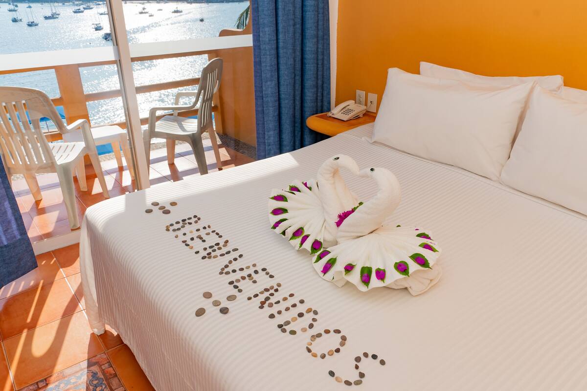 A hotel room with a bed, a balcony with chairs, and a view of boats on the water.
