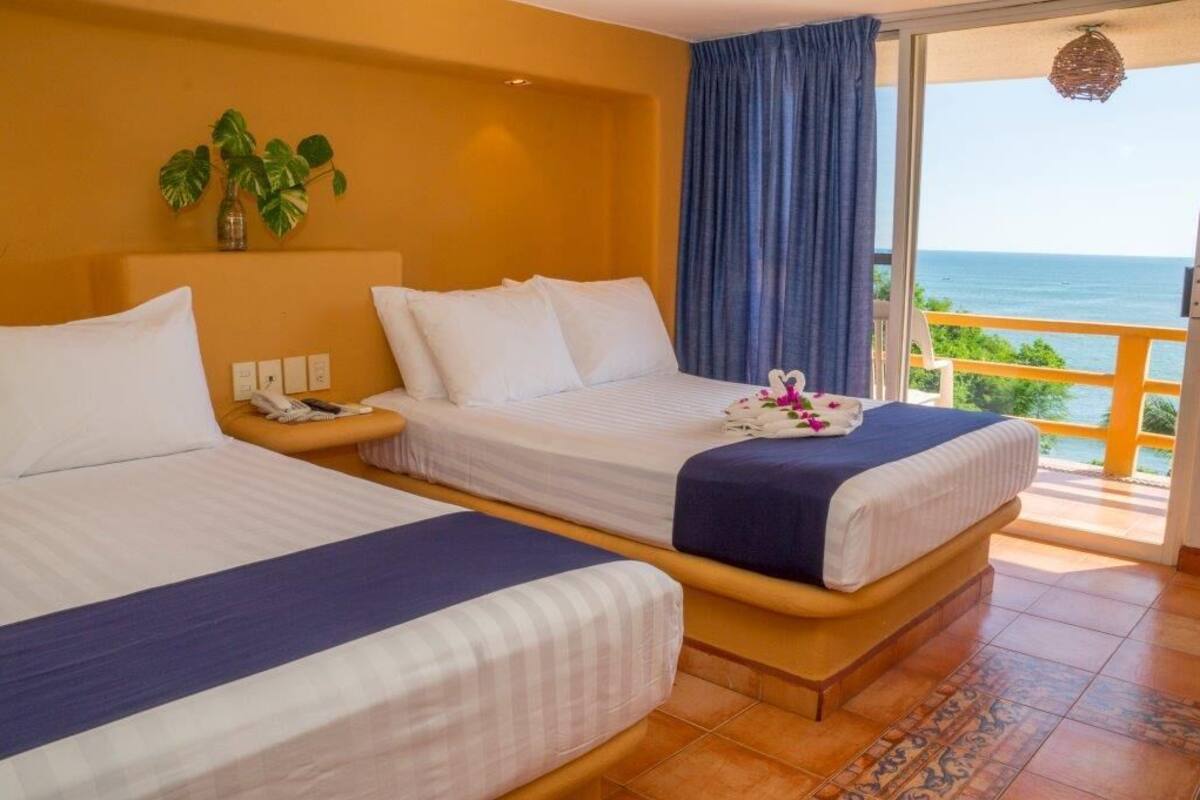 A hotel room with two beds, a balcony with a view of the sea, and a plant on the wall.